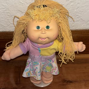 May include: A vintage Cabbage Patch Kids doll with blonde yarn hair and green eyes. The doll wears a two-tone pink and yellow top and a patterned skirt with a white base and colorful geometric designs. The doll is standing on a white base.