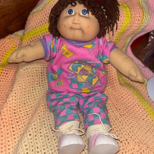 Pode incluir: Uma boneca Cabbage Patch Kid vintage com cabelo de fio castanho e olhos azuis. A boneca usa uma roupa estampada em rosa e verde-azulado com calças a condizer e tênis brancos com listras rosa. Um laço vermelho adorna o cabelo da boneca.