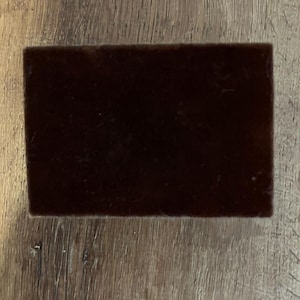Può includere: Un pezzo di tessuto rettangolare di colore marrone scuro con una trama liscia.