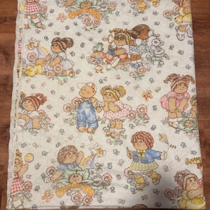 May include: A folded, vintage-style blanket featuring a repeating pattern of cartoon children, flowers, and butterflies. The blanket has a white background with a yellow border. The children are dressed in various outfits, and the overall design is whimsical.
