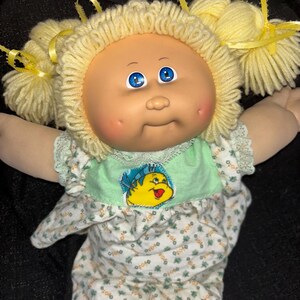 K&ouml;nnte beinhalten: Eine Vintage-Puppe der Cabbage Patch Kids mit blondem Garnhaar in Z&ouml;pfen, die mit gelben B&auml;ndern gebunden sind. Die Puppe hat blaue Augen, rosige Wangen und tr&auml;gt ein Outfit mit gr&uuml;nem und wei&szlig;em Blumenmuster mit einem Cartoon-Motiv.