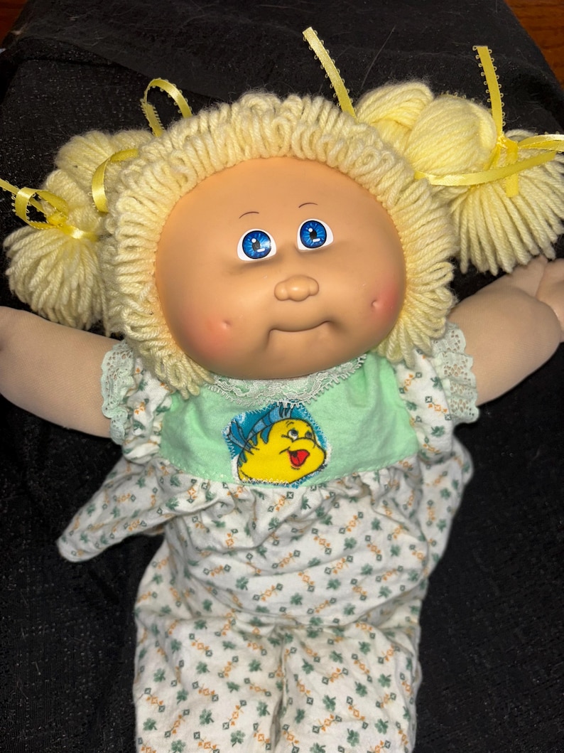 K&ouml;nnte beinhalten: Eine Vintage-Puppe der Cabbage Patch Kids mit blondem Garnhaar in Z&ouml;pfen, die mit gelben B&auml;ndern gebunden sind. Die Puppe hat blaue Augen, rosige Wangen und tr&auml;gt ein Outfit mit gr&uuml;nem und wei&szlig;em Blumenmuster mit einem Cartoon-Motiv.