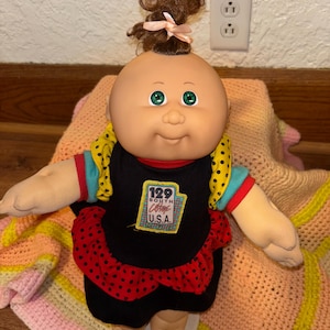 Peut inclure: Une poupée Cabbage Patch Kids vintage avec une tête chauve, des yeux verts et une petite touffe de cheveux bruns attachée avec un ruban rose. La poupée porte une robe noire à pois rouges et un haut jaune et bleu. La poupée est sur une base blanche.
