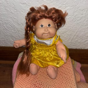 Może przedstawiać: Zabytkowa lalka Cabbage Patch Kid z rudymi włosami w warkoczu. Lalka ma na sobie żółtą satynową sukienkę z białym kołnierzem. Siedzi na różowo-brzoskwiniowym, szydełkowym kocu.
