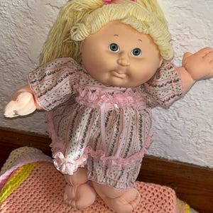 Puede incluir: Una muñeca vintage con cabello rubio de hilo y ojos azules. La muñeca viste un atuendo rosa con estampado floral y ribetes con volantes. Un lazo rosa adorna el cabello de la muñeca. Un juguete clásico.