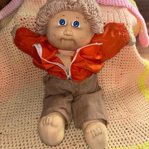 Pode incluir: Uma boneca Cabbage Patch Kid vintage com olhos azuis, vestindo uma jaqueta laranja e calças marrons. A boneca tem um penteado de malha e está deitada em um cobertor de crochê com cores rosa, amarelo e pêssego.