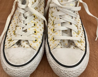 Puede incluir: Zapatillas Converse blancas de lona con lunares dorados. Las zapatillas tienen cordones negros y una suela de goma negra. El logotipo de Converse es visible en el lateral de las zapatillas.