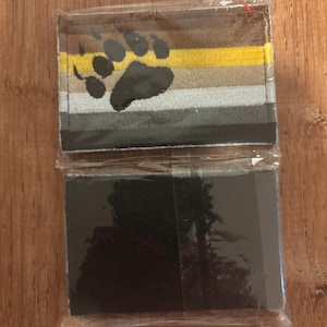 May include: Two patches in plastic packaging. The top patch features a bear paw print over a horizontal striped design in brown, yellow, gray, and black. The bottom patch is solid black.