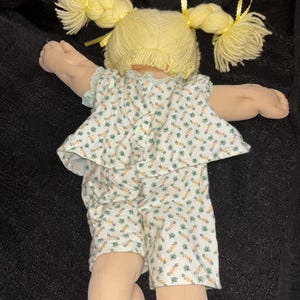 K&ouml;nnte beinhalten: Eine Vintage-Puppe mit blondem Garnhaar, das zu Z&ouml;pfen gestylt und mit gelben B&auml;ndern verziert ist. Die Puppe tr&auml;gt ein wei&szlig;es Outfit mit gr&uuml;nem Blumenmuster. Die Arme und Beine der Puppe sind ausgestreckt.