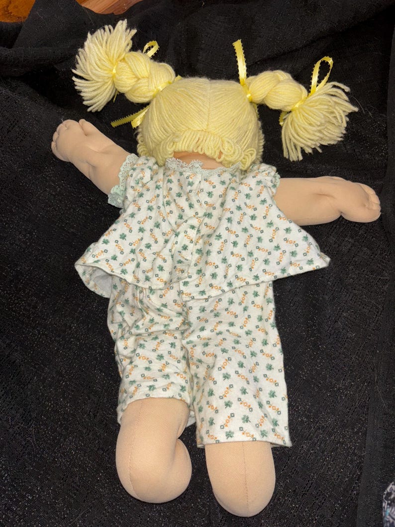 K&ouml;nnte beinhalten: Eine Vintage-Puppe mit blondem Garnhaar, das zu Z&ouml;pfen gestylt und mit gelben B&auml;ndern verziert ist. Die Puppe tr&auml;gt ein wei&szlig;es Outfit mit gr&uuml;nem Blumenmuster. Die Arme und Beine der Puppe sind ausgestreckt.