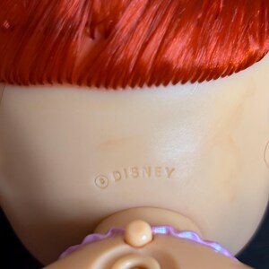 May include: Back view of a red-haired doll with a purple ribbon around her neck. The doll has a light-colored skin tone and a Disney copyright symbol with the word "DISNEY" printed on her back.