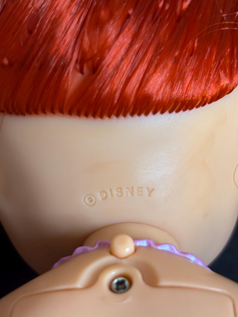 May include: Back view of a red-haired doll with a purple ribbon around her neck. The doll has a light-colored skin tone and a Disney copyright symbol with the word "DISNEY" printed on her back.