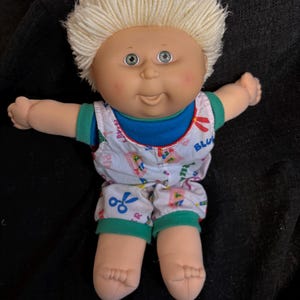Pode incluir: Uma boneca Cabbage Patch Kid com cabelo de lã loiro, olhos verdes e bochechas rosadas. A boneca está vestindo uma roupa azul e verde com um padrão branco de tesouras, réguas e outros materiais escolares.