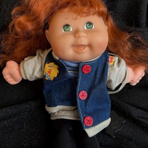 May include: A Cabbage Patch Kid doll with red hair, green eyes, and a denim vest with red buttons. The doll is wearing a white shirt with a blue stripe and black pants.