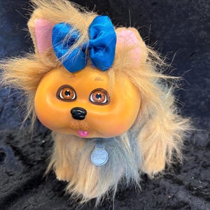 Puede incluir: Un perro de peluche vintage con una cara redonda naranja, ojos marrones y una nariz negra. Tiene un lazo azul, orejas rosas y una etiqueta que dice "CBLC". El pelaje del perro es una mezcla de marrón, gris y azul.