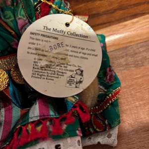 Op de afbeelding: Een close-up van een pop uit The Muffy Collection. De pop draagt een groene, rode en witte jurk met gouden accenten. Op het label staat "The Muffy Collection" en bevat veiligheidsvoorschriften.