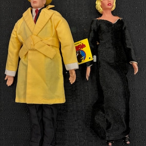 May include: Two vintage dolls. One doll is dressed in a yellow trench coat, hat, and black pants. The other doll is wearing a black dress. A yellow tag with text is attached to the black dress.