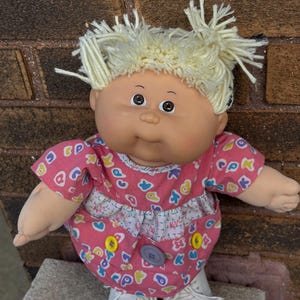 May include: A vintage Cabbage Patch Kid doll with blonde yarn hair and a pink dress decorated with colorful shapes. The doll has a round face, brown eyes, and is wearing white trainers. The dress has yellow and blue buttons.