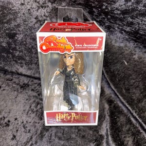 May include: A boxed vinyl collectible figure of a character from the Harry Potter series. The figure is in a clear plastic case with a red and white box featuring the text "Harry Potter" and "Rock Candy". The figure has brown hair and is wearing a black outfit.
