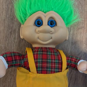 Puede incluir: Una muñeca troll vintage con cabello verde brillante y ojos azules. La muñeca viste una camisa a cuadros roja y verde y un mono amarillo. La muñeca tiene una expresión sonriente y manos de tela blanca.
