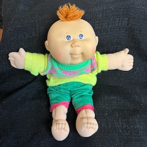 Könnte beinhalten: Eine Vintage Cabbage Patch Kid-Puppe mit kahlem Kopf, blauen Augen und einem Büschel orangefarbenem Haar. Die Puppe trägt einen neongrünen und rosa Pullover, grüne Shorts mit rosa Besatz und beige Schuhe. Die Puppe sitzt mit ausgestreckten Armen.