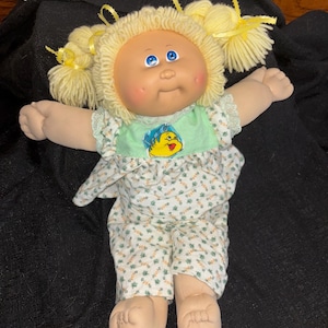 May include: A vintage Cabbage Patch Kids doll with blonde yarn hair in pigtails, decorated with yellow ribbons. The doll is dressed in a green and white floral print outfit with a cartoon graphic on the bodice. It has blue eyes and rosy cheeks.