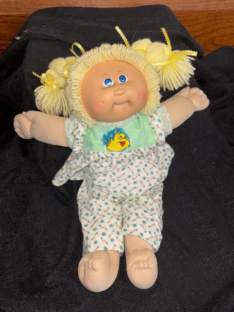 K&ouml;nnte beinhalten: Eine Vintage-Cabbage-Patch-Kids-Puppe mit blondem Garnhaar, das zu Z&ouml;pfen geflochten und mit gelben B&auml;ndern verziert ist. Die Puppe tr&auml;gt ein Outfit mit gr&uuml;nem und wei&szlig;em Blumenmuster und einem Cartoon-Motiv auf dem Oberteil. Sie hat blaue Augen und rosige Wangen.