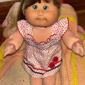 Puede incluir: Una muñeca vintage con ojos verdes, cabello castaño y una diadema morada. La muñeca viste un vestido blanco con estampados florales rojos, ribetes rojos y un diseño de cerezas. La muñeca está sobre una manta multicolor.