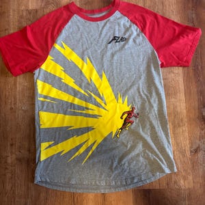 May include: A grey t-shirt with red raglan sleeves featuring a large yellow lightning bolt graphic on the front. The shirt displays a small black Flash logo on the upper chest and a graphic of the character The Flash running to the right.