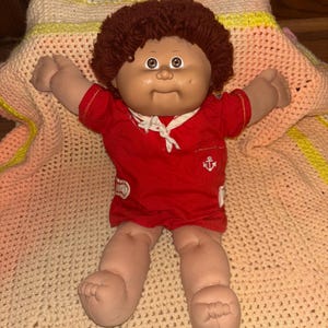 May include: A vintage Cabbage Patch Kid doll with brown hair and a red sailor outfit. The doll has a tan face and arms, and is lying on a colorful crocheted blanket. The red outfit has a white collar and an anchor emblem.
