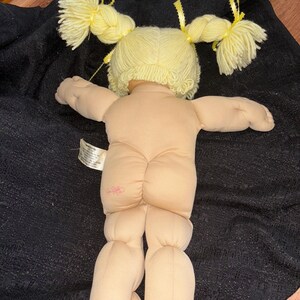 K&ouml;nnte beinhalten: Eine Vintage-Puppe mit hellem Hautton, gelbem Garnhaar, das zu zwei Z&ouml;pfen gestylt ist, und einem rosa Blumenemblem auf der R&uuml;ckseite. Die Puppe liegt auf einer schwarzen Oberfl&auml;che. Die Arme und Beine der Puppe sind ausgestreckt.
