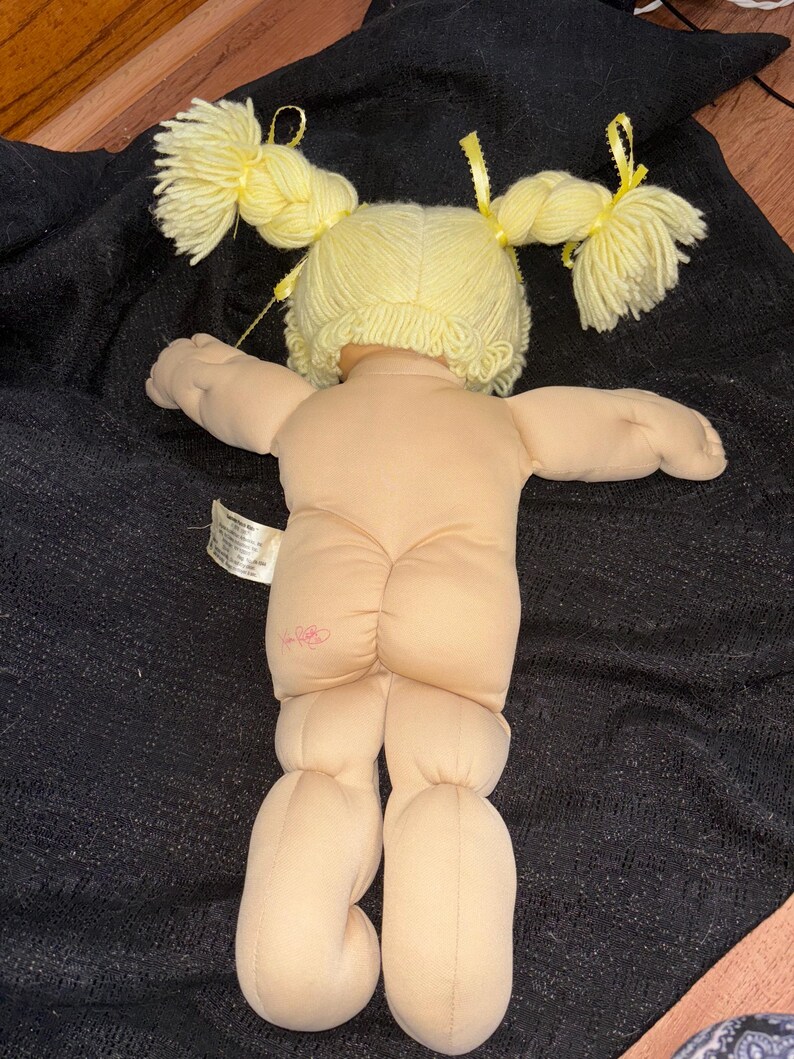 K&ouml;nnte beinhalten: Eine Vintage-Puppe mit hellem Hautton, gelbem Garnhaar, das zu zwei Z&ouml;pfen gestylt ist, und einem rosa Blumenemblem auf der R&uuml;ckseite. Die Puppe liegt auf einer schwarzen Oberfl&auml;che. Die Arme und Beine der Puppe sind ausgestreckt.