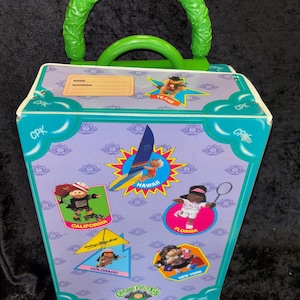 May include: A vintage Cabbage Patch Kids travel case with a green handle and trim. The case features illustrations of Cabbage Patch Kids representing various U.S. states, including Texas, California, Hawaii, Florida, Colorado, and New York. The case has a purple background.