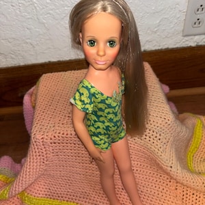 May include: A vintage doll with long blonde hair and green eyes, wearing a green and blue patterned romper. The doll is posed on a pink, yellow, and white crocheted blanket. The doll's face has pink blush and lipstick.