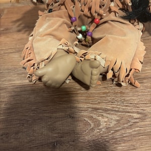 May include: A Native American doll with tan suede clothing and fringed details. The doll has beaded accents and tan-coloured feet. The doll is sitting on a wooden surface.