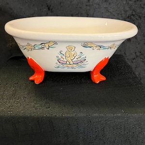 May include: A white ceramic bathtub with orange claw feet. The tub features a whimsical design of a bear on a raft, surrounded by fish and other aquatic elements. The rim of the tub is decorated with swimming fish.