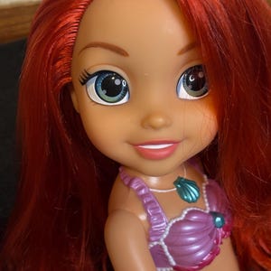 May include: A red-haired doll with green eyes and a purple seashell top. The doll is wearing a necklace with a seashell pendant.