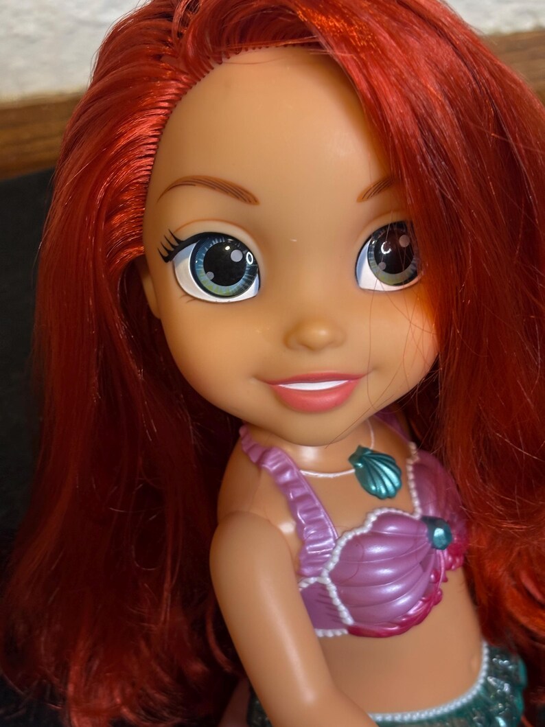 May include: A red-haired doll with green eyes and a purple seashell top. The doll is wearing a necklace with a seashell pendant.