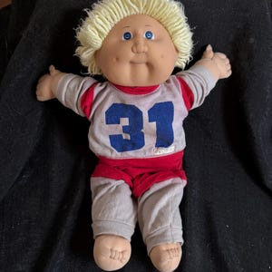 Puede incluir: Una muñeca Cabbage Patch Kid con cabello rubio y ojos azules que lleva una camiseta gris y roja con el número 31 en la parte delantera.