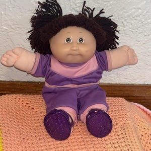 Puede incluir: Una muñeca Cabbage Patch Kid vintage con cabello de hilo marrón peinado en coletas. La muñeca viste un atuendo morado y rosa con zapatos de purpurina morados a juego. Está sentada sobre una manta de ganchillo color melocotón.