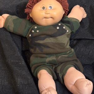 May include: A Cabbage Patch Kid doll with brown yarn hair wearing a camouflage print outfit with silver studs. The doll is lying on a dark gray surface.