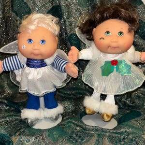 Può includere: Due bambole angelo con capelli biondi e castani, occhi blu e verdi e guance rosee. Le bambole indossano abiti bianchi e blu con ali e poggiano su basi argentate e dorate. Una bambola ha una stella sulla guancia, l'altra un motivo a bacche di agrifoglio.