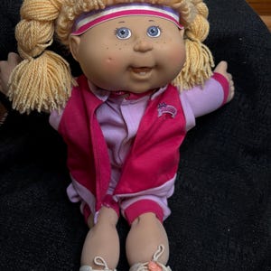 May include: A vintage Cabbage Patch Kids doll with blonde yarn hair styled in pigtails. The doll has blue eyes, freckles, and is wearing a pink and purple athletic outfit with white sneakers. The doll's outfit includes a pink vest with a logo.