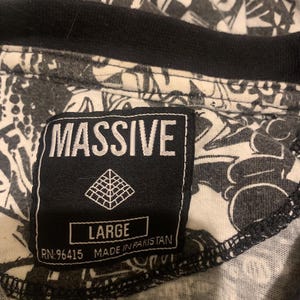 Puede incluir: Una camiseta gráfica de manga corta en blanco y negro con una etiqueta negra que dice "MASSIVE" en letras blancas. La etiqueta también tiene un logotipo de pirámide blanca y el texto "LARGE RN-96415 MADE IN PAKISTAN".