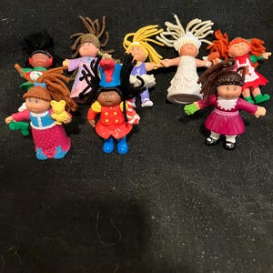 May include: A collection of eight Cabbage Patch Kids dolls, each with unique outfits and hairstyles. The dolls are made of plastic and have a variety of colors, including red, blue, green, yellow, white, and brown.