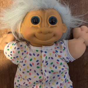 Può includere: Una bambola Troll vintage con capelli blu e grandi occhi blu. La bambola indossa una tutina bianca con pois colorati. Ha un viso sorridente e un paio di orecchie da coniglio blu.