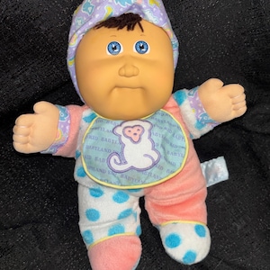 May include: A Cabbage Patch Kid doll with a soft, plush body. The doll has a peach-coloured face, blue eyes, and a purple bonnet with "CPK" on it. It wears a bib with a monkey graphic and a pink and blue outfit.