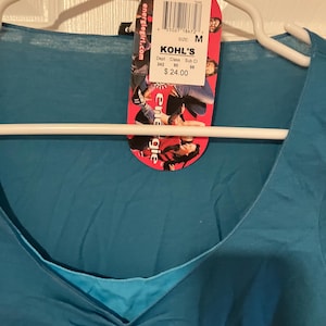 May include: A teal blue v-neck t-shirt with a white tag attached. The tag has the text "KOHL'S" and "$24.00" printed on it. The tag also has a barcode and the text "SIZE: M" printed on it.