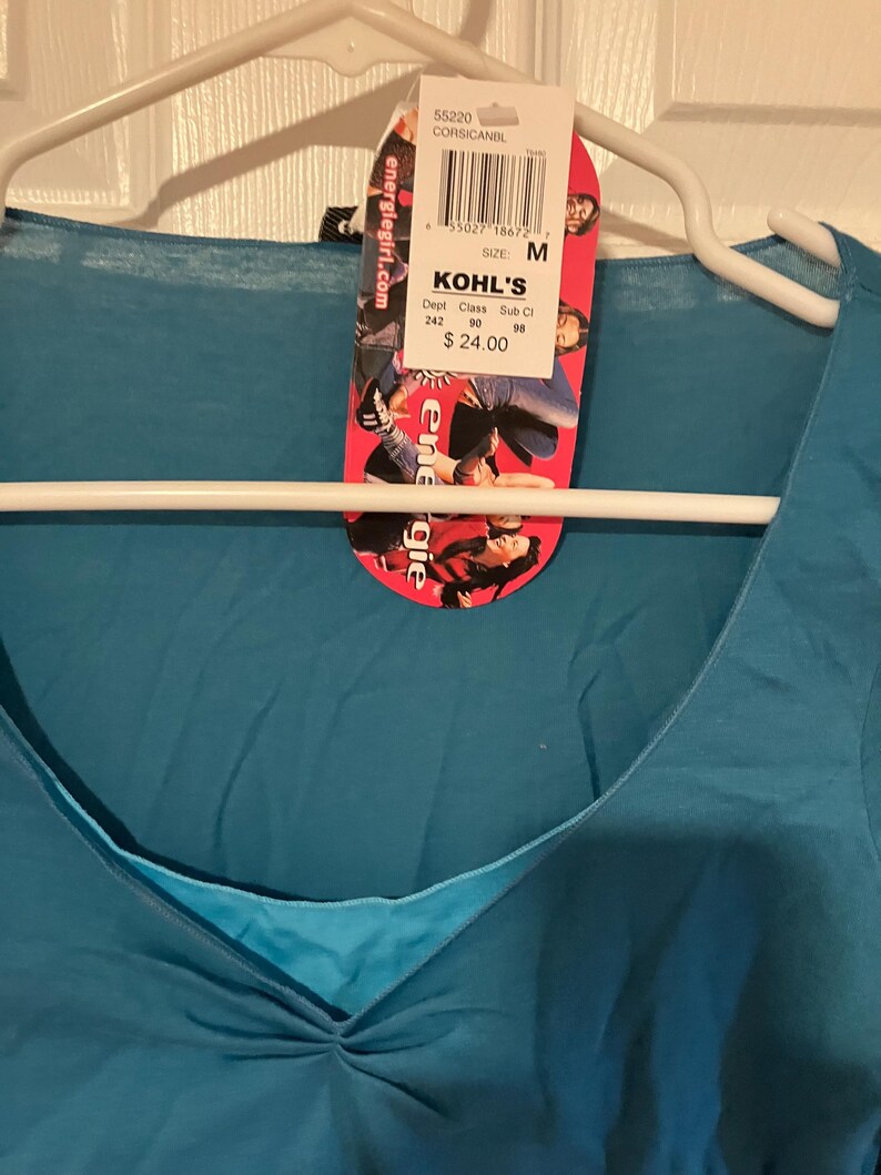 May include: A teal blue v-neck t-shirt with a white tag attached. The tag has the text "KOHL'S" and "$24.00" printed on it. The tag also has a barcode and the text "SIZE: M" printed on it.