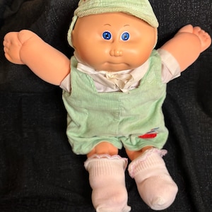 May include: A vintage Cabbage Patch Kid doll with blue eyes, wearing a light green corduroy outfit, a matching cap with a red cross, and white socks with pink trim. The doll has its arms outstretched.